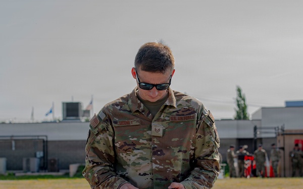 A U.S. Air Force service member in OCP (Operational Camouflage Pattern) uniform stands at a table outdoors, appearing to handle or inspect a magazine or small equipment item. Several items are laid out on the table in front of him, suggesting a gear inspection, equipment check, or timed competition event. In the background, a group of other military personnel are gathered near a building on what appears to be a military installation.