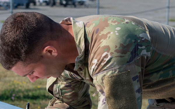 A male military service member in MultiCam/OCP camouflage uniform is leaning over a metal table outdoors, writing on a document attached to a clipboard. The individual appears focused on completing paperwork, possibly a form or sign-in sheet. The setting appears to be an outdoor military installation or training area.