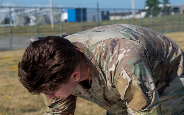 A military service member in OCP (Operational Camouflage Pattern) uniform is leaning over a metal table outdoors, writing on paper with a blue pen. The setting appears to be a military installation or training area, with a fenced compound and open grassy field visible in the background. The individual appears to be completing paperwork or documentation.