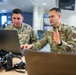 Two U.S. Air Force personnel in OCP (Operational Camouflage Pattern) uniforms are collaborating at a table while reviewing content on a laptop computer. One airman is gesturing with his hand while appearing to explain or discuss something on the screen. Professional camera equipment is visible on the table, suggesting a public affairs or media-related work context.