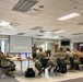 A military briefing or workshop is taking place in what appears to be a conference or dining facility. A presenter in camouflage uniform stands before a projection screen displaying 'The Strategic Lens' presentation, addressing a group of approximately 12-15 military personnel seated in a semicircular arrangement. The event appears to be an Air Force visual storytelling and strategic communications workshop.