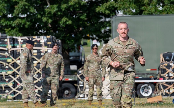 A group of military personnel in OCP (Operational Camouflage Pattern) uniforms are gathered in a grassy field near military equipment and vehicles. One soldier in the foreground is jogging toward the camera, while three others stand in the background near stacked cargo pallets and a military truck. The setting appears to be a logistics or staging area, likely during a training exercise or operational deployment.