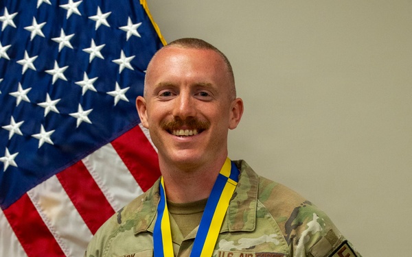 A U.S. Air Force Security Forces member named 'Slick' poses proudly in front of an American flag wearing OCP (Operational Camouflage Pattern) uniform. He is displaying a large championship-style belt and medal on a blue and yellow lanyard, both bearing the 'Defensor Fortis' Security Forces insignia. The individual appears to be receiving recognition for a competition or achievement within the Security Forces community.