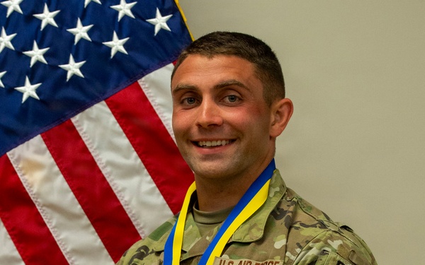 A U.S. Air Force service member in OCP (Operational Camouflage Pattern) uniform poses in front of an American flag while wearing a blue and yellow medal ribbon around their neck. The individual displays multiple patches including 'SF' (Security Forces), 'RAVEN', and a National Guard patch on the left sleeve. The photo appears to be an official or semi-official recognition portrait.
