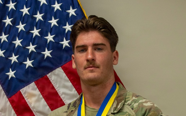 A U.S. Air Force service member named Casey stands at attention in front of an American flag wearing OCP (Operational Camouflage Pattern) uniform. He is wearing a bronze medal on a blue and yellow ribbon lanyard, likely from a competition or ceremony. Patches on his right sleeve indicate he is assigned to a Security Forces (SF) unit within the Air National Guard.