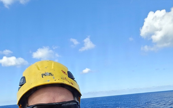 A man takes a selfie outdoors over open ocean, wearing professional safety equipment including a yellow Petzl helmet and a 3M DBI-SALA fall arrest harness. The background shows a deep blue ocean horizon under a partly cloudy sky, suggesting he is positioned at height on a marine structure such as a ship mast, offshore platform, or wind turbine. His equipment indicates he is engaged in a professional working-at-height activity in an offshore environment.