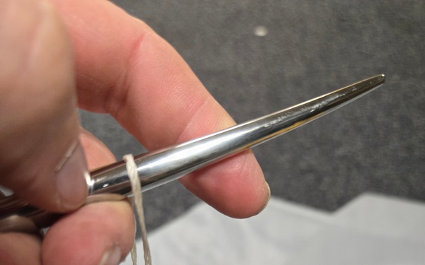 A hand is using a large metal sailmaker's or upholstery needle threaded with white twine to hand-stitch along the edge of a gray fabric or vinyl material. Several completed stitches are visible along the seam, suggesting a repair or construction task in progress. Yellow and blue clamps are visible in the lower portion of the image, holding the material in place.