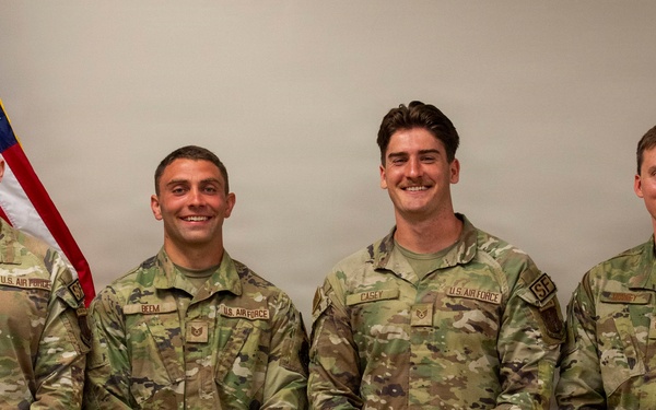 Four U.S. Air Force personnel in OCP (Operational Camouflage Pattern) uniforms are posed for a formal group photo. They are standing in front of an American flag and what appears to be a unit or service flag. Name tapes visible on their uniforms include 'SLICK', 'BEEM', 'CASEY', and a partially visible fourth name.