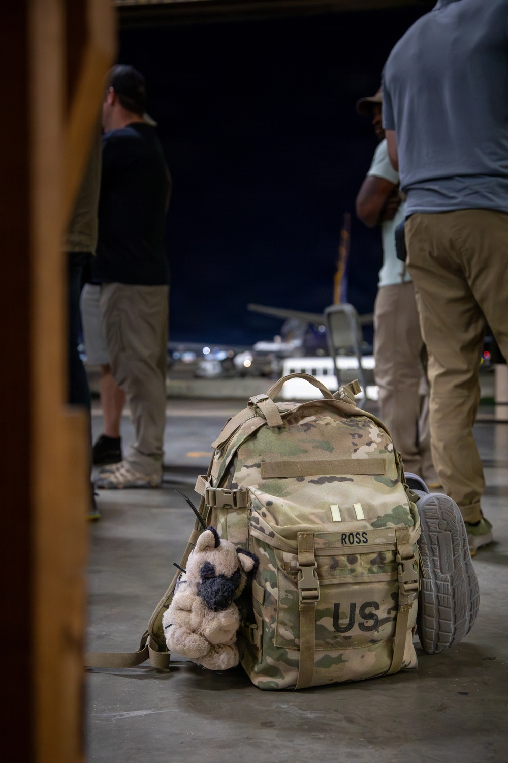 A US military multicam-pattern backpack labeled 'ROSS' sits on a concrete floor at what appears to be a military airfield or hangar at night. A small stuffed animal toy (appearing to be a pug or similar dog) is attached to the side of the pack, adding a personal touch. Several individuals in civilian attire stand nearby with aircraft visible in the background.