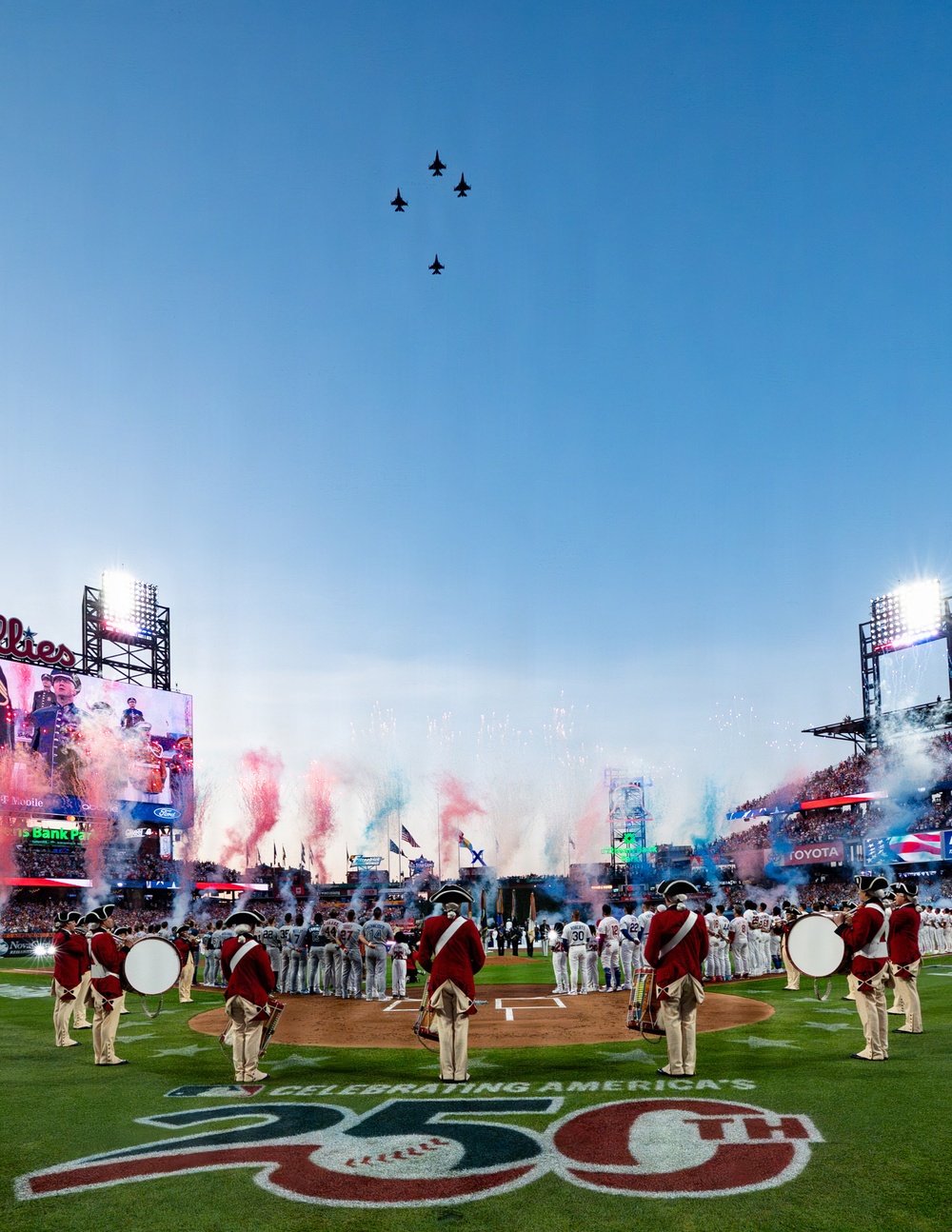 A grand ceremonial pre-game event at Citizens Bank Park, home of the Philadelphia Phillies, celebrating America's 250th anniversary. The scene features a military flyover of four jet aircraft, red/white/blue pyrotechnic smoke effects, Revolutionary War-era costumed drummers in red coats, and baseball players lined up on the field. The stadium is packed with fans and illuminated by large video boards and stadium lights.