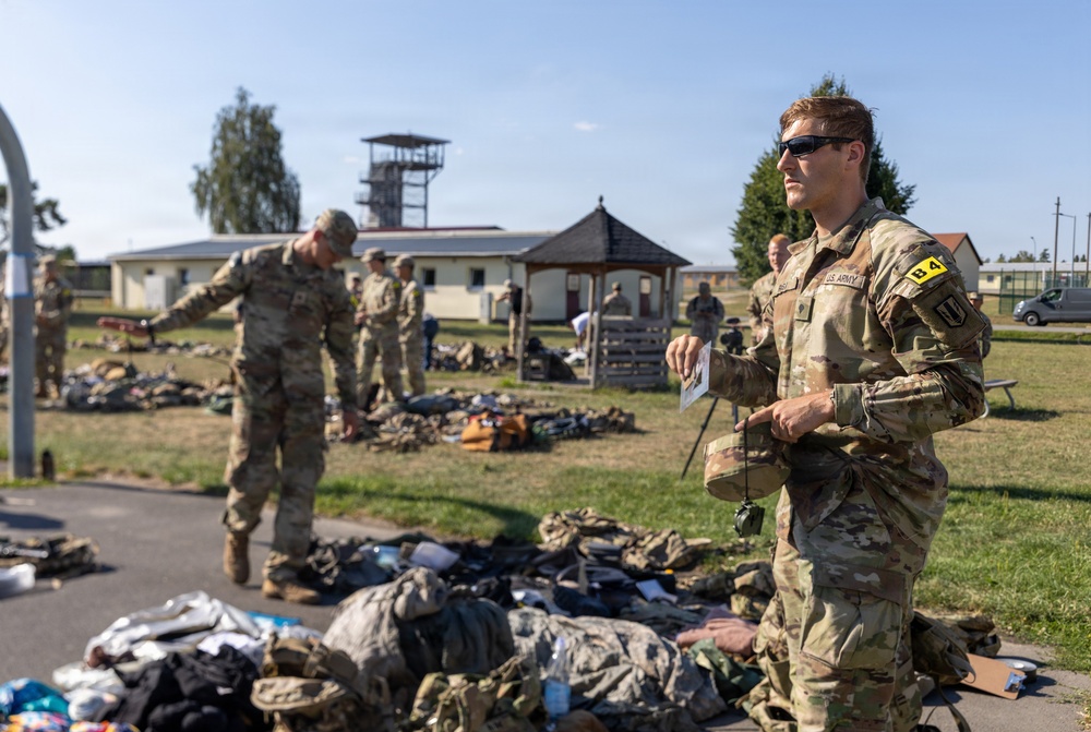 U.S. Army soldiers in OCP (Operational Camouflage Pattern) uniforms are conducting what appears to be a gear inspection or equipment layout on a military installation. Multiple soldiers are standing among piles of military equipment and personal gear spread across a grassy area. The setting appears to be a European military base based on the architectural style of the buildings visible in the background.