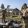 USAREUR-AF Best Squad Competition Day Zero Layout U.S. Army soldiers in OCP (Operational Camouflage Pattern) uniforms are conducting what appears to be a gear inspection or equipment layout on a military installation. Multiple soldiers are standing among piles of military equipment and personal gear spread across a grassy area. The setting appears to be a European military base based on the architectural style of the buildings visible in the background.