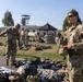 U.S. Army soldiers in OCP (Operational Camouflage Pattern) uniforms are conducting what appears to be a gear inspection or equipment layout on a military installation. Multiple soldiers are standing among piles of military equipment and personal gear spread across a grassy area. The setting appears to be a European military base based on the architectural style of the buildings visible in the background.