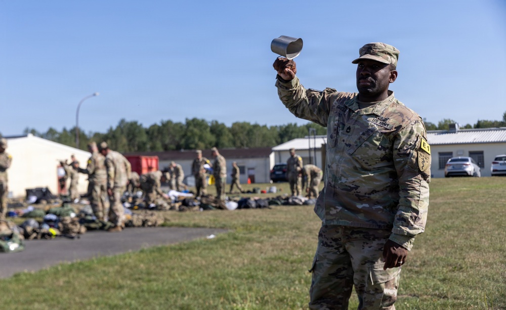 A U.S. Army soldier in OCP (Operational Camouflage Pattern) uniform holds up a metal cup or canteen cup with one raised arm in the foreground. Behind him, multiple soldiers are gathered in an outdoor area near military buildings, with equipment and gear spread on the ground, suggesting a field inspection, layout drill, or equipment accountability formation. The setting appears to be a military installation or training area.