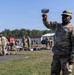 A U.S. Army soldier in OCP (Operational Camouflage Pattern) uniform holds up a metal cup or canteen cup with one raised arm in the foreground. Behind him, multiple soldiers are gathered in an outdoor area near military buildings, with equipment and gear spread on the ground, suggesting a field inspection, layout drill, or equipment accountability formation. The setting appears to be a military installation or training area.