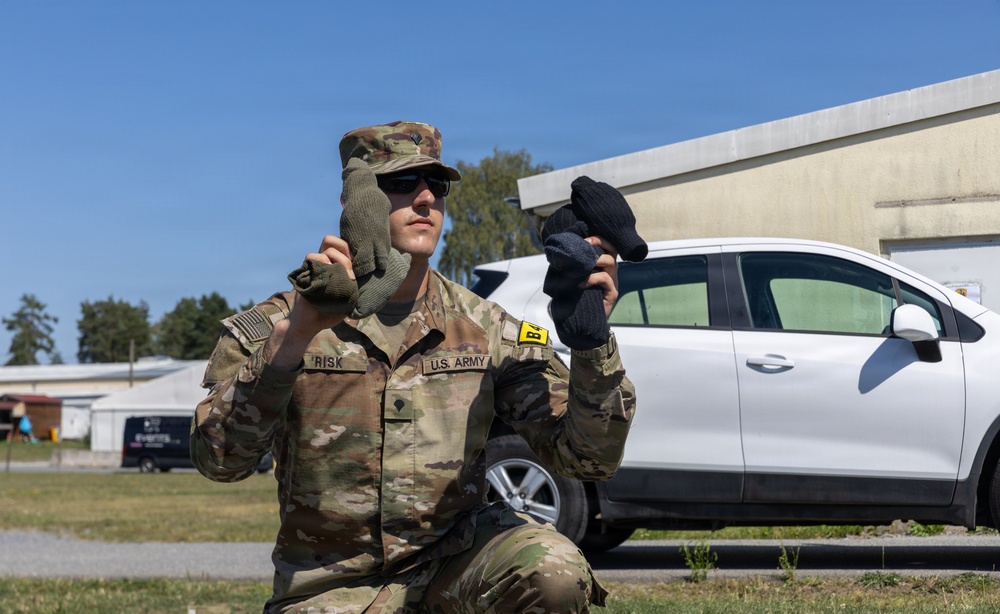 A U.S. Army soldier wearing OCP (Operational Camouflage Pattern) uniform, identified by the name tape 'RISK', is kneeling outdoors while holding up two different gloves for comparison — one olive/tan tactical glove on the left hand and one black glove in the right hand. The soldier is wearing sunglasses and an OCP patrol cap. The setting appears to be a military installation or base, with a white civilian vehicle and a concrete building visible in the background.