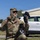 USAREUR-AF Best Squad Competition Day Zero Layout A U.S. Army soldier wearing OCP (Operational Camouflage Pattern) uniform, identified by the name tape 'RISK', is kneeling outdoors while holding up two different gloves for comparison — one olive/tan tactical glove on the left hand and one black glove in the right hand. The soldier is wearing sunglasses and an OCP patrol cap. The setting appears to be a military installation or base, with a white civilian vehicle and a concrete building visible in the background.