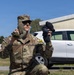 A U.S. Army soldier wearing OCP (Operational Camouflage Pattern) uniform, identified by the name tape 'RISK', is kneeling outdoors while holding up two different gloves for comparison — one olive/tan tactical glove on the left hand and one black glove in the right hand. The soldier is wearing sunglasses and an OCP patrol cap. The setting appears to be a military installation or base, with a white civilian vehicle and a concrete building visible in the background.