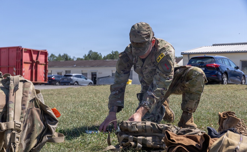 A U.S. Army soldier in OCP (Operational Camouflage Pattern) uniform is crouched on a grass field, inspecting or organizing tactical gear and equipment laid out on the ground. The soldier appears to be at a military installation or training area, with buildings, vehicles, and a red shipping container visible in the background. The scene suggests pre-mission preparation or equipment inspection.