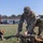 USAREUR-AF Best Squad Competition Day Zero Layout A U.S. Army soldier in OCP (Operational Camouflage Pattern) uniform is crouched on a grass field, inspecting or organizing tactical gear and equipment laid out on the ground. The soldier appears to be at a military installation or training area, with buildings, vehicles, and a red shipping container visible in the background. The scene suggests pre-mission preparation or equipment inspection.
