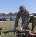 A U.S. Army soldier in OCP (Operational Camouflage Pattern) uniform is crouched on a grass field, inspecting or organizing tactical gear and equipment laid out on the ground. The soldier appears to be at a military installation or training area, with buildings, vehicles, and a red shipping container visible in the background. The scene suggests pre-mission preparation or equipment inspection.