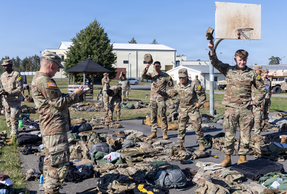 A group of U.S. Army soldiers in OCP (Operational Camouflage Pattern) uniforms are conducting what appears to be a field equipment inspection or layout drill on a paved outdoor area of a military installation. Soldiers have spread their gear, packs, and equipment on the ground while a non-commissioned officer reviews a clipboard, likely conducting an inventory check. The scene takes place near a basketball court on what appears to be a European military base, indicated by building styles and a Red Cross vehicle visible in the background.