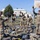 USAREUR-AF Best Squad Competition Day Zero Layout A group of U.S. Army soldiers in OCP (Operational Camouflage Pattern) uniforms are conducting what appears to be a field equipment inspection or layout drill on a paved outdoor area of a military installation. Soldiers have spread their gear, packs, and equipment on the ground while a non-commissioned officer reviews a clipboard, likely conducting an inventory check. The scene takes place near a basketball court on what appears to be a European military base, indicated by building styles and a Red Cross vehicle visible in the background.