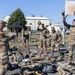 A group of U.S. Army soldiers in OCP (Operational Camouflage Pattern) uniforms are conducting what appears to be a field equipment inspection or layout drill on a paved outdoor area of a military installation. Soldiers have spread their gear, packs, and equipment on the ground while a non-commissioned officer reviews a clipboard, likely conducting an inventory check. The scene takes place near a basketball court on what appears to be a European military base, indicated by building styles and a Red Cross vehicle visible in the background.