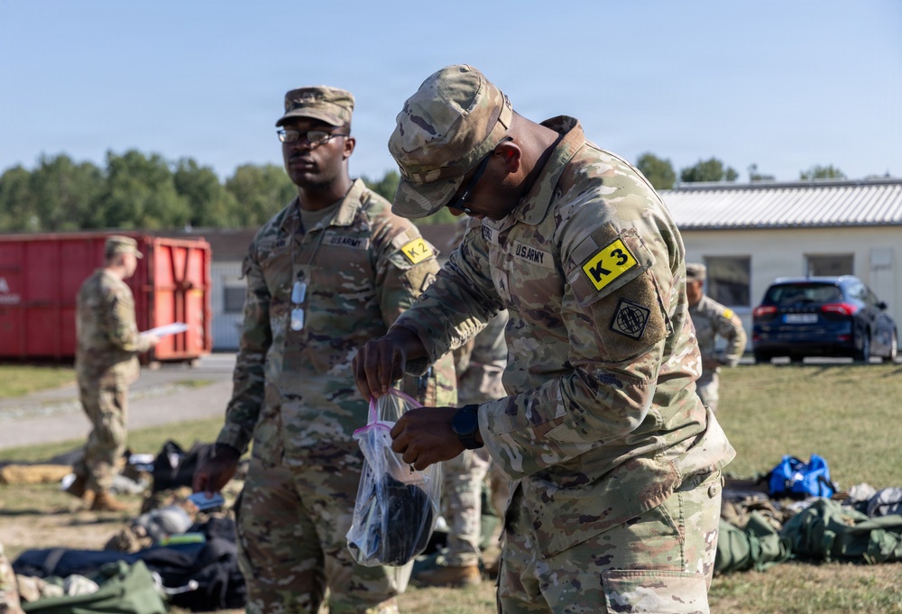 Two U.S. Army soldiers in OCP (Operational Camouflage Pattern) uniforms are engaged in what appears to be an equipment inspection or inventory activity outdoors. The foreground soldier wears a yellow 'K3' patch and is examining items from a clear plastic bag, while the background soldier wears a 'K2' patch. The setting appears to be a military installation or training area with gear spread on the ground.