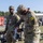 USAREUR-AF Best Squad Competition Day Zero Layout Two U.S. Army soldiers in OCP (Operational Camouflage Pattern) uniforms are engaged in what appears to be an equipment inspection or inventory activity outdoors. The foreground soldier wears a yellow 'K3' patch and is examining items from a clear plastic bag, while the background soldier wears a 'K2' patch. The setting appears to be a military installation or training area with gear spread on the ground.