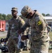 Two U.S. Army soldiers in OCP (Operational Camouflage Pattern) uniforms are engaged in what appears to be an equipment inspection or inventory activity outdoors. The foreground soldier wears a yellow 'K3' patch and is examining items from a clear plastic bag, while the background soldier wears a 'K2' patch. The setting appears to be a military installation or training area with gear spread on the ground.