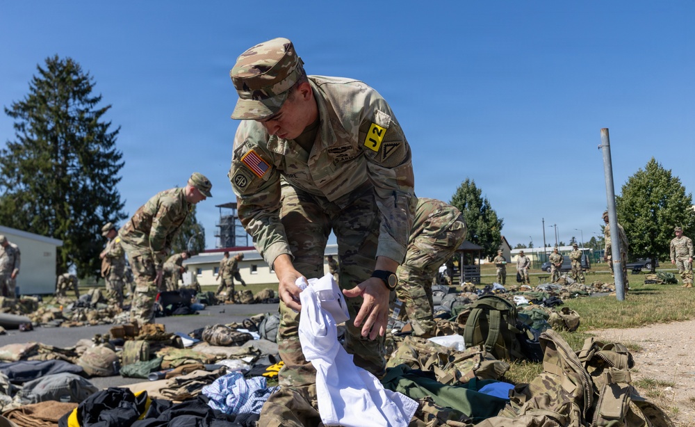 Multiple U.S. Army soldiers in OCP (Operational Camouflage Pattern) uniforms are conducting what appears to be a layout inspection or equipment accountability check outdoors on a military installation. Gear, bags, clothing items, and tactical equipment are spread across the ground in organized rows. The foreground soldier wears a yellow 'J2' staff identifier tab and an American flag patch, suggesting an intelligence staff role.