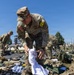 Multiple U.S. Army soldiers in OCP (Operational Camouflage Pattern) uniforms are conducting what appears to be a layout inspection or equipment accountability check outdoors on a military installation. Gear, bags, clothing items, and tactical equipment are spread across the ground in organized rows. The foreground soldier wears a yellow 'J2' staff identifier tab and an American flag patch, suggesting an intelligence staff role.