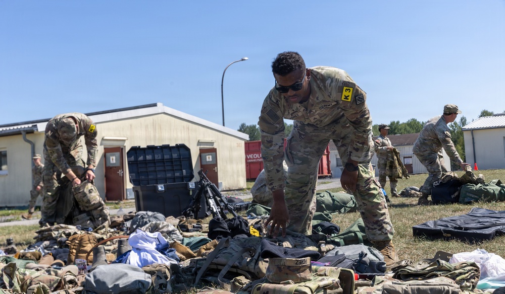 Multiple U.S. Army soldiers in OCP (Operational Camouflage Pattern) uniforms are sorting through large piles of military gear and equipment outdoors on a grassy area. The soldiers appear to be conducting a layout inspection or equipment accountability check, a common military administrative procedure. The setting appears to be a military installation or training area in a temperate region.