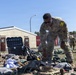 Multiple U.S. Army soldiers in OCP (Operational Camouflage Pattern) uniforms are sorting through large piles of military gear and equipment outdoors on a grassy area. The soldiers appear to be conducting a layout inspection or equipment accountability check, a common military administrative procedure. The setting appears to be a military installation or training area in a temperate region.