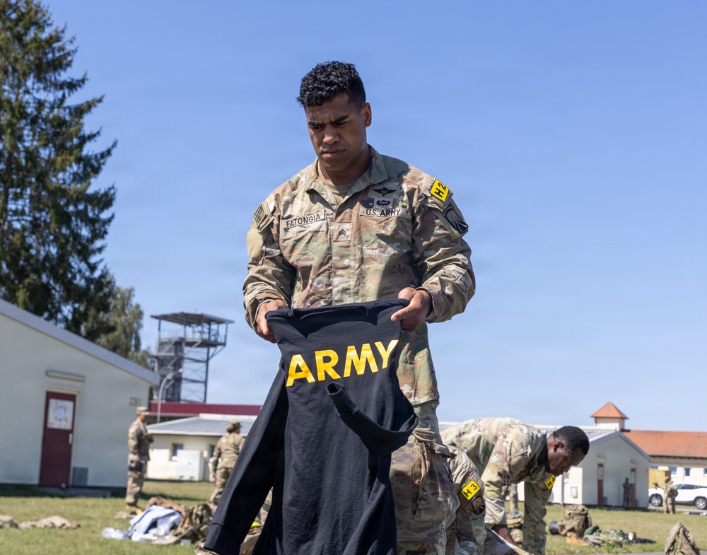 A U.S. Army Sergeant named Fatongia is holding up a black Army physical training shirt outdoors on a military installation. Multiple soldiers in OCP (Operational Camouflage Pattern) uniforms are visible in the background engaged in similar activities. The setting appears to be a field inspection or uniform layout exercise on a sunny day.