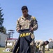 A U.S. Army Sergeant named Fatongia is holding up a black Army physical training shirt outdoors on a military installation. Multiple soldiers in OCP (Operational Camouflage Pattern) uniforms are visible in the background engaged in similar activities. The setting appears to be a field inspection or uniform layout exercise on a sunny day.