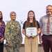 Four individuals are posing together at what appears to be an award ceremony or recognition event. A young woman in the center is holding a 'Certificate of Appreciation' made out to 'Margaret Maggie Delany.' The group includes a civilian woman, a U.S. Navy service member in camouflage uniform with the name tag 'RAZA,' and a bearded civilian man.