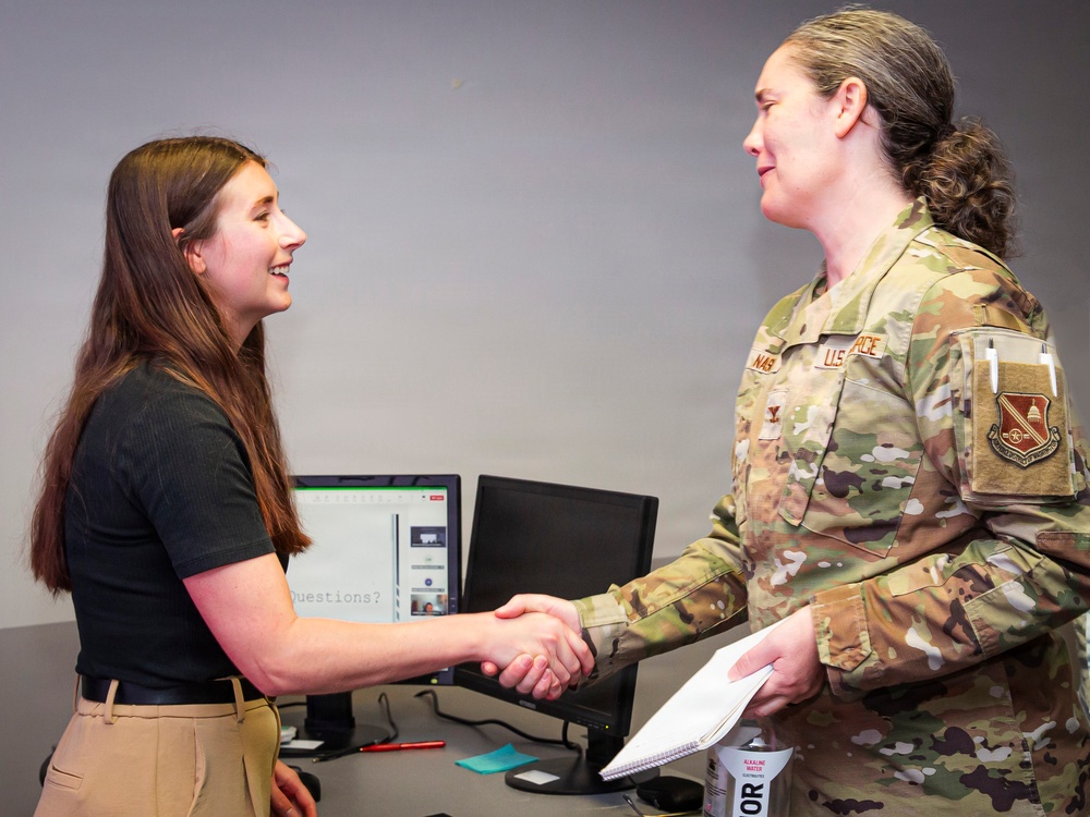 Two women are shaking hands across a desk in what appears to be an office or briefing room setting. One woman is in civilian attire while the other is wearing a U.S. Air Force OCP (Operational Camouflage Pattern) uniform. The interaction appears cordial and professional, possibly following a presentation or meeting.