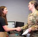 Two women are shaking hands across a desk in what appears to be an office or briefing room setting. One woman is in civilian attire while the other is wearing a U.S. Air Force OCP (Operational Camouflage Pattern) uniform. The interaction appears cordial and professional, possibly following a presentation or meeting.