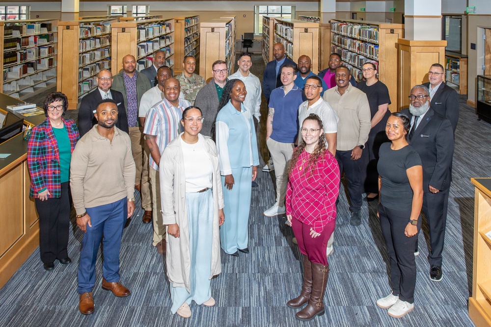 A large group of approximately 25 adults is posed for a formal group photograph inside a well-lit library. The individuals are arranged in multiple rows between tall wooden bookshelves filled with books. The group appears diverse in gender, age, and ethnicity, dressed in business casual to semi-formal attire.