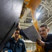 Two personnel in dark blue coveralls/uniforms are performing maintenance work on a piece of yellow industrial or military equipment. One individual is physically working on the machinery while wearing blue gloves, and the other is referencing a printed document or checklist. The setting appears to be an indoor industrial or military vessel/hangar bay environment.