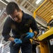 A U.S. Navy sailor in dark blue coveralls is working on a mechanical component while standing next to a yellow 36V electric forklift or ground support equipment. The sailor is wearing blue nitrile gloves and appears to be inspecting or servicing a small mechanical part using a tool. The setting appears to be an indoor hangar or maintenance facility.
