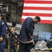 U.S. Navy personnel are conducting fueling or fluid transfer operations aboard what appears to be a naval vessel's hangar bay. Two sailors are prominently featured in the foreground, one managing a large fuel hose while wearing protective eyewear and gloves, and another looking upward while monitoring the operation. A large American flag is displayed prominently in the background.