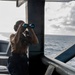 A military or naval personnel member stands on the deck of a warship, using binoculars to scan the open ocean. The individual is wearing a tan/khaki shirt, navy blue trousers, and a helmet or headset, suggesting an active watch or lookout duty. The ship's metal superstructure frames the scene, with choppy open ocean and overcast skies visible in the background.