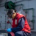 A female service member in naval working uniform is crouched on what appears to be the deck of a military vessel, performing a cleaning or maintenance task. She is wearing a red safety vest over blue coveralls, protective goggles, and blue nitrile gloves, and is working with a pink rag and a small tool near a metal bucket. The background shows gray ship bulkhead with green piping, valves, a fire hose reel, and a watertight door.