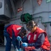 Three personnel in matching red and navy blue uniforms are working on the deck of a naval vessel, engaged in a cleaning or maintenance task involving buckets and brushes. They are wearing protective equipment including safety goggles and blue nitrile gloves. The setting is clearly a ship's exterior deck, with firefighting and safety station signage visible in the background.