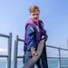 A young male sailor in US Navy blue coveralls and a purple flotation/work vest is handling a large black hose or line on the deck of a naval vessel. He is wearing black work gloves and combat boots, and appears to be exerting effort while managing the equipment. The background shows open water and a distant shoreline under a clear blue sky.