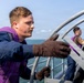 Two personnel wearing purple vests over flight deck uniforms are working on the deck of a naval vessel at sea. They appear to be handling or operating deck equipment, possibly related to aircraft handling or mooring operations. The setting is an open ocean environment under partly cloudy skies.