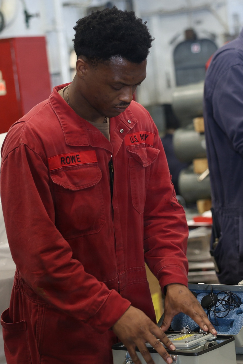 A U.S. Navy sailor named Rowe, wearing red coveralls, is working with equipment in what appears to be a shipboard workspace or maintenance area. He is focused on handling items in a foam-lined case or tray. The setting appears to be an interior compartment of a naval vessel.